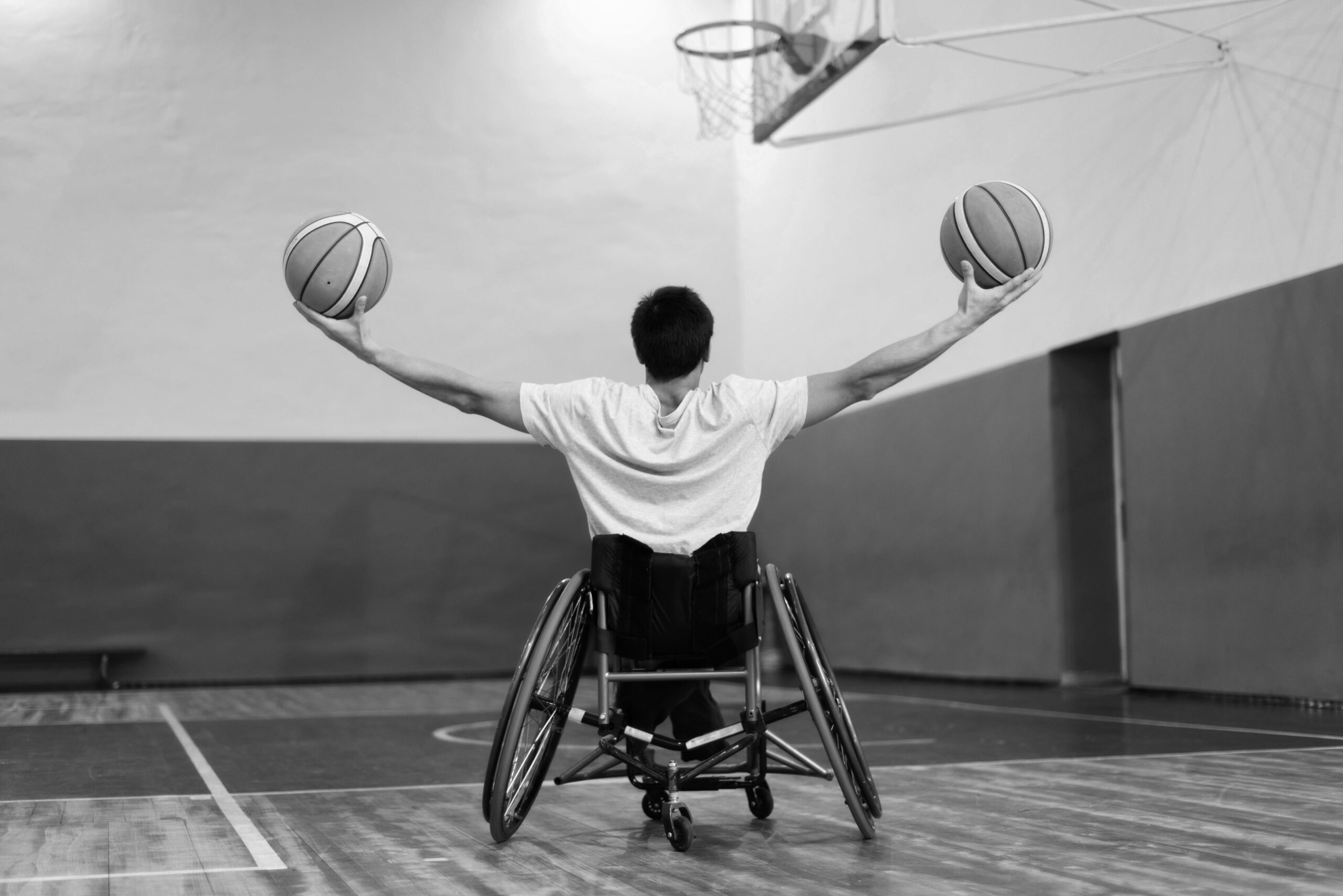 Un homme en fauteuil de dos, bras écartés tenant un ballon de basket dans chaque main.